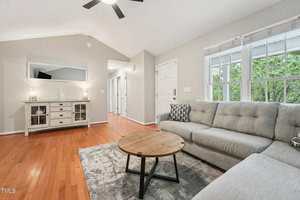 A living room with hardwood floors, a gray sectional sofa, a round wooden coffee table, a patterned rug, a white sideboard with lamps, a wall mirror, and large windows showing greenery outside.