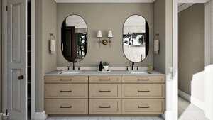A modern bathroom features a double sink vanity with light wood cabinets, two oval mirrors, a wall-mounted light fixture, and neutral-toned walls. A potted plant and soap dispensers sit on the countertop.