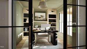 A modern home office with glass doors, a large desk with a computer monitor, bookshelves on either side, a painting on the wall, a white rug, and large windows letting in natural light.