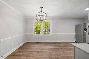 A dining area with light-colored walls, wooden floor, a large window with a view of greenery, a modern spherical chandelier, and part of a stainless steel refrigerator visible on the right.