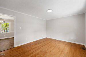 An empty room with light gray walls, white trim, and a hardwood floor. A round ceiling light is on the white ceiling. An open doorway leads to another room with a window and similar flooring.