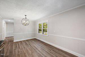 A bright, empty room with light gray walls, white trim, wood-style flooring, a modern circular chandelier, a large window, and a hallway visible to the left.
