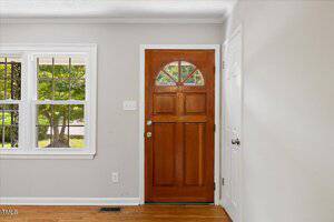 A wooden front door with a semi-circular window at the top, next to a white interior closet door and a window with a view of greenery outside; hardwood flooring and neutral-colored walls.