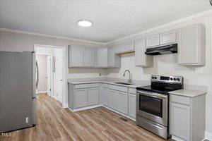 A modern kitchen with light gray cabinets, stainless steel appliances, an electric stove with a range hood, a double-door refrigerator, and wood-look flooring. Walls are light-colored and the ceiling has a round light fixture.