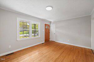 A bright, empty room with light gray walls, a hardwood floor, a large window with white trim, and a wooden front door. A round ceiling light is centered on the white ceiling.