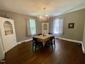 A dining room with wooden floors, two windows with sheer curtains, a small table covered with a colorful cloth, four chairs, built-in corner cabinets, and a ceiling chandelier.
