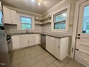 A kitchen with white cabinets, patterned tile backsplash, gray tile floor, stainless steel appliances, open shelves, a window above the sink, and a white door with a window.