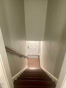 A view from the top of a staircase leading down to a closed white door with a rug in front. The walls are beige, and there is a wooden handrail on the left side.