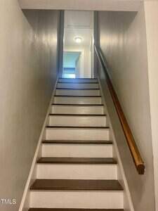 A staircase with white risers and brown treads leads up to a hallway with a ceiling light. The walls are painted beige, and there is a wooden handrail on the right side.