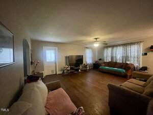 A living room with hardwood floors, three couches, a TV on a stand, two windows with patterned curtains, a ceiling fan with light, and a door to the left of the TV.