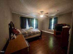 A bedroom with dark wood floors, a bed with plaid bedding, a wooden dresser with water bottles, a ceiling fan with light, two windows with dark curtains, and a wooden entertainment center.