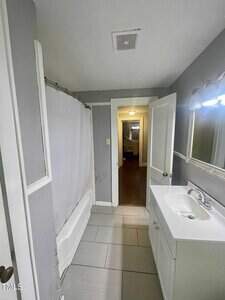 A small bathroom with gray walls, a white bathtub with a shower curtain, a white vanity with a sink and mirror, tile flooring, and an open door leading to a hallway with hardwood floors.