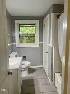 A small bathroom with gray walls and tile floor, a white vanity with sink, a toilet, a window above the toilet, and a shower with a white curtain. There is visible water damage on the wall near the tub.