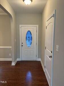 A white front door with an oval stained glass window is set in a beige hallway with wood flooring. A closed white closet door is to the right, and an arched doorway is on the left side of the image.