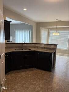 A modern kitchen with dark wood cabinets, granite countertops, a double sink, and a dishwasher. The kitchen opens to a bright dining area with large windows and a hanging light fixture.