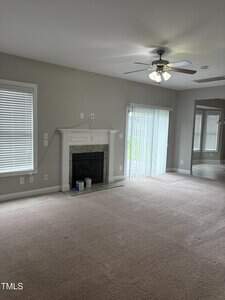A carpeted living room with light gray walls, a ceiling fan with lights, a fireplace with a white mantel, two paint cans on the hearth, large windows with blinds, and glass sliding doors leading outside.