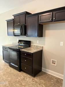 A kitchen with dark wood cabinets, a black electric stove, a matching microwave, granite countertops, and an open space with a washer/dryer hookup on the right wall. The floor is tiled.