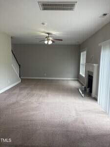 A spacious, empty living room with beige carpet, light gray walls, a ceiling fan with lights, a staircase on the left, a fireplace on the right, and large windows with vertical blinds letting in natural light.