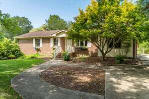 Single-story brick house with a small porch, white trim, and a brown roof. A curved concrete walkway leads to the entrance. Mature trees and landscaping surround the house, with a clear sky in the background.