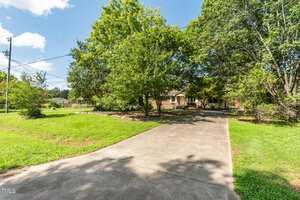 A concrete driveway leads to a single-story brick house partially obscured by large green trees, with a well-maintained lawn on either side and a clear blue sky overhead.