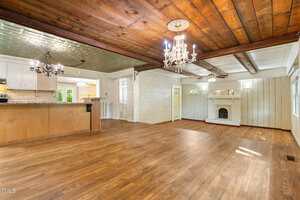 Spacious open living area features wood floors, exposed wood beams, a chandelier, a white fireplace, and an adjoining kitchen with light wood cabinetry and tile backsplash. Natural light enters through several windows.