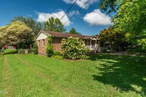A single-story brick house with a covered front porch, surrounded by green grass, shrubs, and trees under a blue sky with scattered clouds. A driveway and a red shed are visible on the left side.