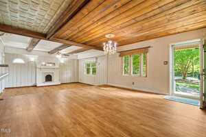 Spacious room with wood flooring, exposed wood-beam ceiling, white paneled walls, a fireplace, a chandelier, and large windows. An open door leads outside to a green yard.