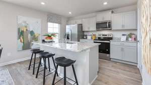 Modern kitchen with light gray cabinets, stainless steel appliances, and a center island with three black stools. Abstract art hangs on the left wall, and a window brings in natural light. Light wood flooring throughout.