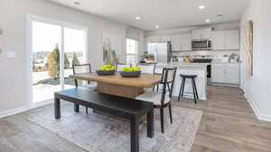 A modern kitchen and dining area features a wooden table with a bench and chairs, bowls of green apples, white cabinets, stainless steel appliances, and a sliding glass door leading outside.