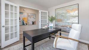 A modern home office features a black desk with a laptop and books, a white upholstered chair, wall art, and double French doors leading to an adjacent hallway with additional abstract artwork.