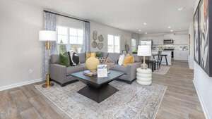 A modern open-concept living room with a gray sectional sofa, yellow and white pillows, a black coffee table, and a floor lamp, adjacent to a white kitchen and dining area with wood-style flooring.