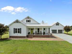 A white, single-story farmhouse at 4715 Uzzle Road, Rougemont, features a covered front porch, double garage, and gravel driveway, all set on a large green lawn beneath a blue sky with scattered clouds.