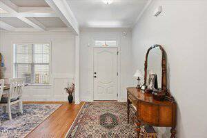 A hallway with a wooden front door, a decorative mirror and table on the right, a patterned rug on the floor, and a dining area with a table and chairs visible to the left.