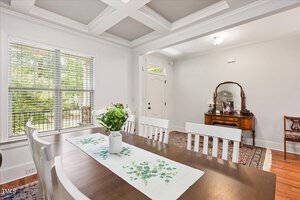 A dining room with a wooden table and white chairs, a floral centerpiece, and a runner. Large window with blinds on the left, a rug, and a vintage wooden sideboard with a mirror against the far wall.
