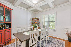 A dining room with a dark wood table, six white chairs, a red and black china cabinet, and a wooden hutch. There is a floral centerpiece on the table, a large window, and a decorative area rug on the hardwood floor.