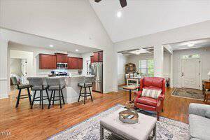 Open-concept living area with hardwood floors, a kitchen with stainless steel appliances and a breakfast bar, red cabinets, and a living space with seating, a rug, and decor. Entryway and additional room visible.