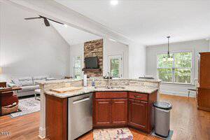 View from a kitchen with a marble countertop, stainless steel dishwasher, and sink, looking into a living room with a stone fireplace and TV, sofa, windows, and hardwood floors throughout the open space.
