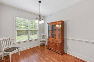 A small dining area with hardwood floors, two white chairs, a wooden china cabinet, white trim, light gray walls, a window with blinds, and a ceiling light fixture.
