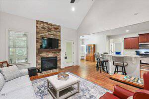 A living room with a stone fireplace, mounted TV, gray sofa, patterned rug, and coffee table. The space opens into a kitchen with bar seating and visible wooden cabinetry and dining area.