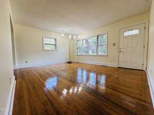 Empty living room with polished hardwood floors, cream-colored walls, three windows, a white front door, and a floor lamp in the corner. Natural light fills the space, creating a bright and clean atmosphere.