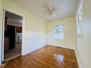 A small, empty room with polished wood floors, cream-colored walls, a ceiling fan, and two windows. An open doorway leads to a kitchen area with visible appliances and wood cabinets.