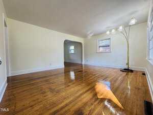 A spacious, empty living room with polished hardwood floors, cream-colored walls, a large arched doorway, multiple windows, and a tall floor lamp with three glass shades. Natural light fills the room.