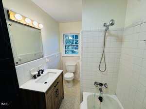 A small bathroom with a mirrored vanity, sink, toilet, and a shower-tub combo with white tile walls. There is a window above the toilet and overhead vanity lights above the mirror.