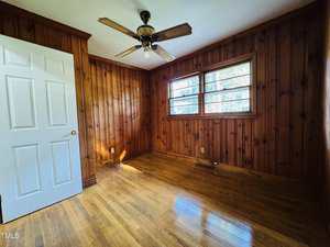 A small empty room with wood-paneled walls, hardwood floors, a ceiling fan with light, a white door, and a window letting in natural light.