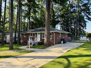 A small, single-story red brick house with a gray roof is surrounded by tall pine trees. The front yard has a curved concrete driveway, a ramp leading to the front door, and green grass.