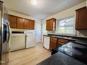 A kitchen with wood cabinets, black countertops, and light wood flooring features a washer, dryer, dishwasher, stove, and stainless steel refrigerator. A window and door are visible on the far wall.
