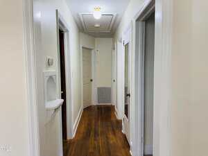 A narrow hallway with white walls and hardwood floors, several open doors on either side, a thermostat and wall niche on the left, and a ceiling light near an attic access door above.