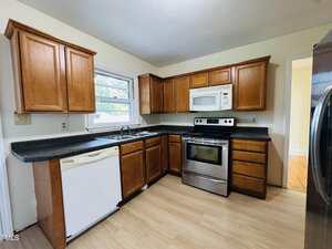 A kitchen with wood cabinets, black countertops, a stainless steel oven and microwave, a white dishwasher, a double sink under a window, and light wood flooring. Walls are painted a light color.