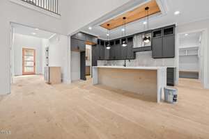 A modern, unfinished kitchen with black cabinets, white countertops, and pendant lights above the island. The floor is bare with construction materials visible, and an open doorway leads to other rooms.
