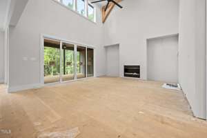 A large, empty living room with unfinished floors, white walls, a built-in fireplace, tall ceiling, ceiling fan, and glass sliding doors leading to a covered outdoor area. Construction materials are on the floor.